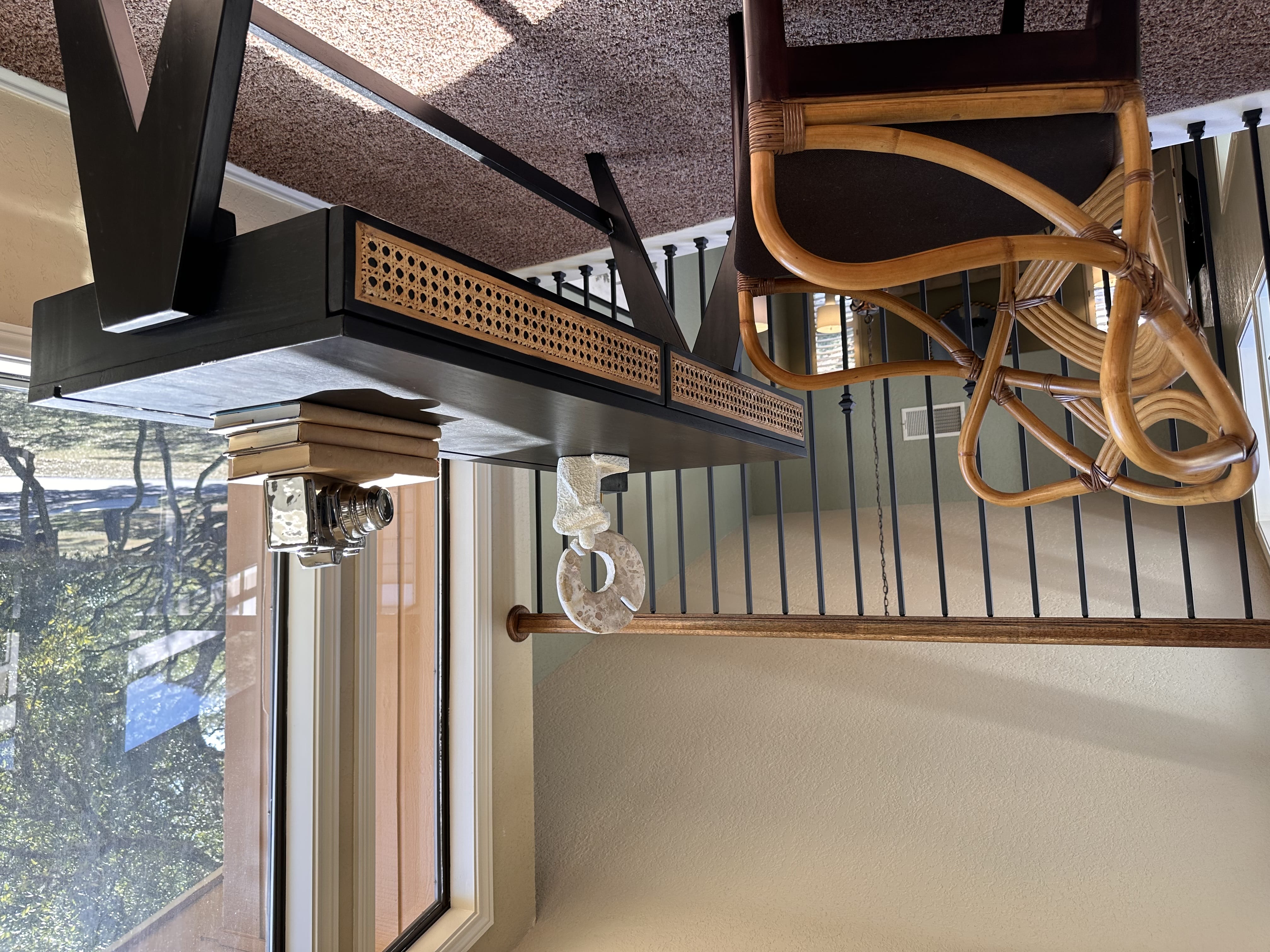 A cozy loft home office setup featuring a black writing desk with woven rattan drawer fronts, paired with an ornate bentwood rattan armchair. The space is situated next to a large window overlooking trees, positioned along an indoor balcony railing.