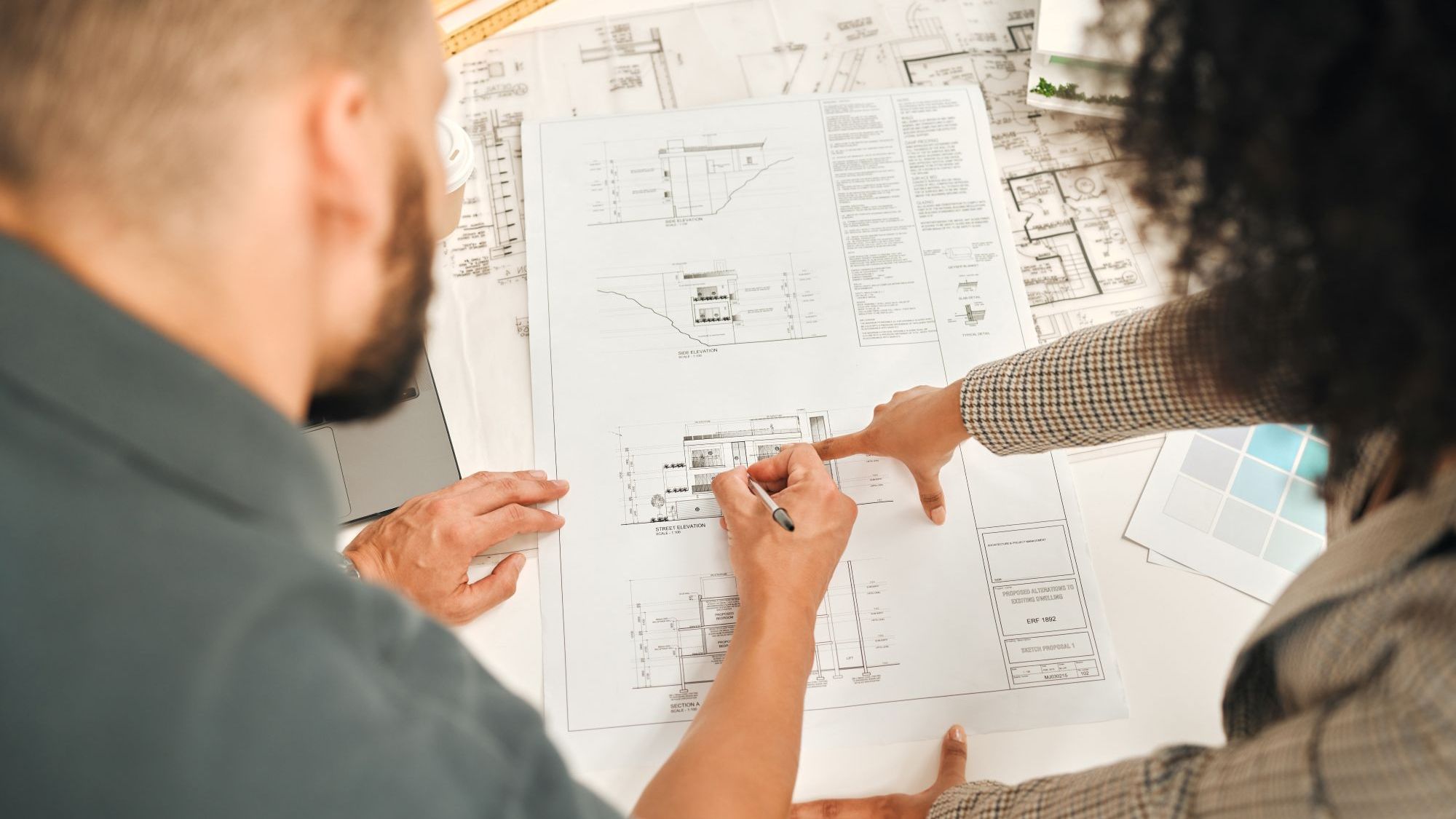 Two professionals reviewing architectural blueprints together, pointing and making notes on detailed building plans.