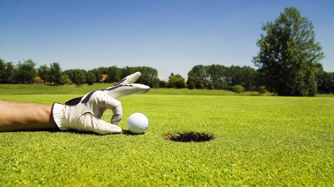 Close-up of a gloved golfer’s hand placing a white golf ball on the green next to the hole under a clear blue sky.
