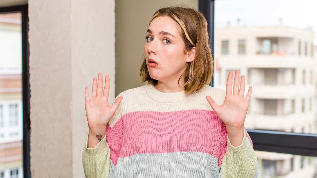 A woman standing indoors with her hands raised in a “not my responsibility” gesture, looking surprised or defensive.