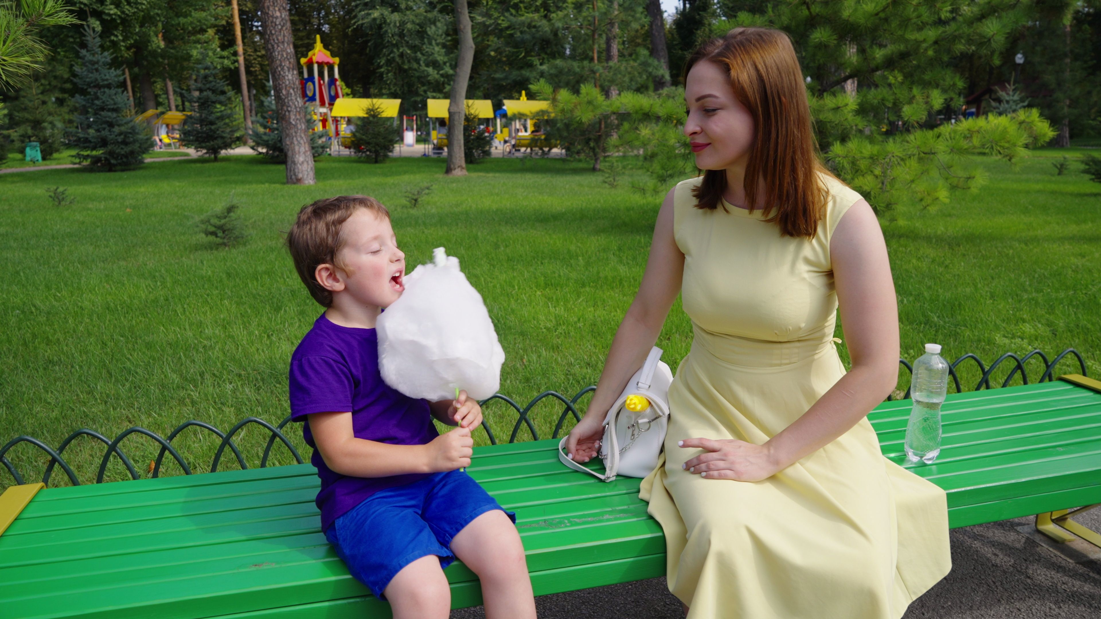 A little boy holds a giant cotton candy cone, while his mother sits nearby.