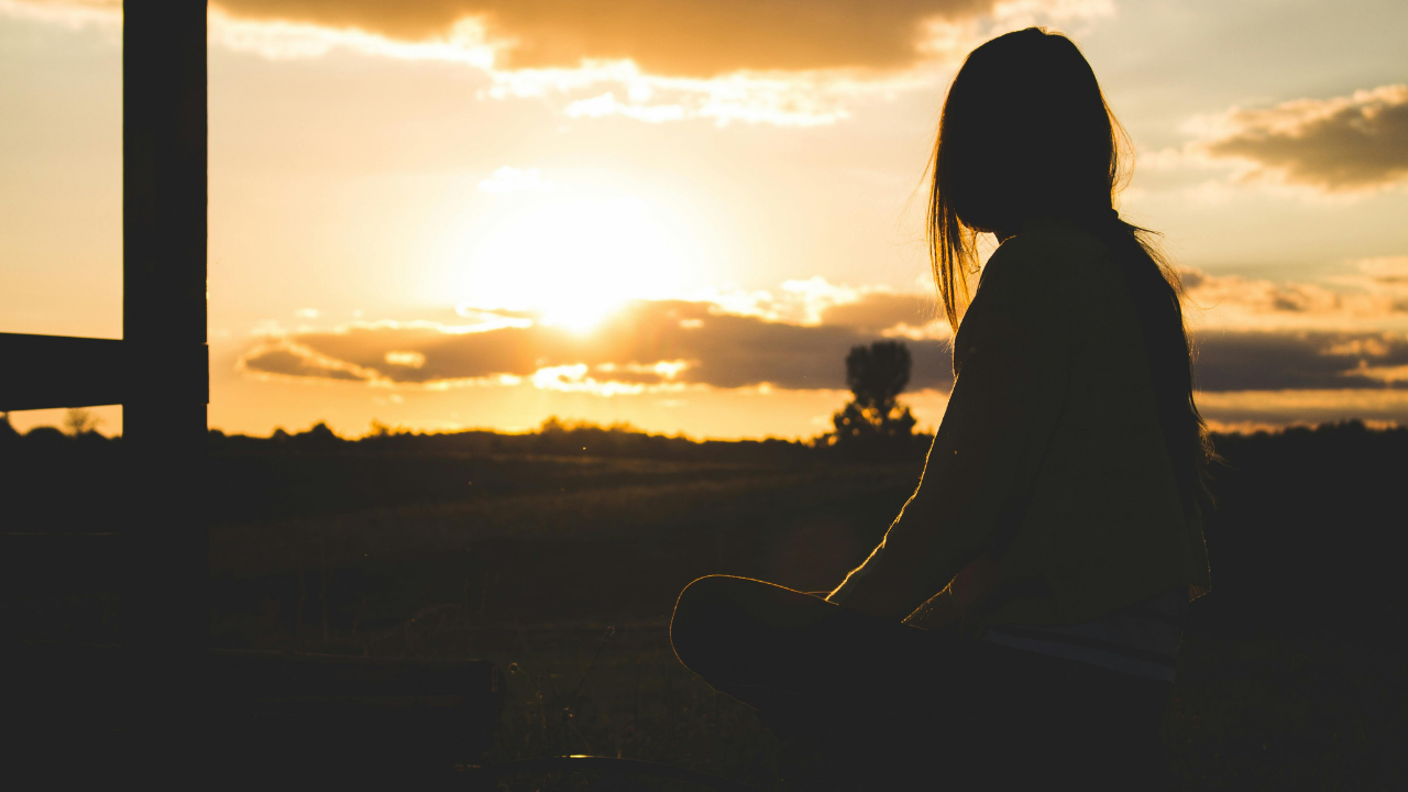 Silhouette of a woman sitting quietly at sunset, reflecting alone while experiencing a subtle sense of emotional distance and inner disconnection.