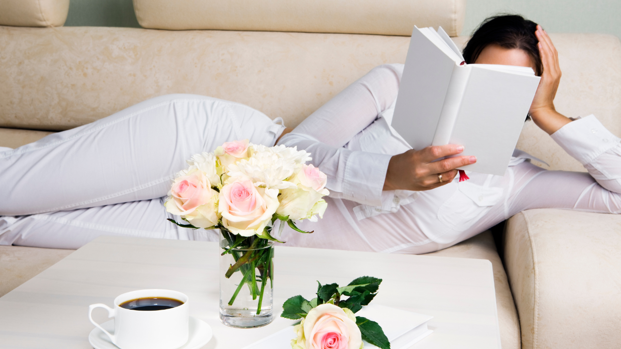 woman lying on side on sofa propped up on left elbow head in hand, reading a book. coffee table in front has roses and a cup of coffee