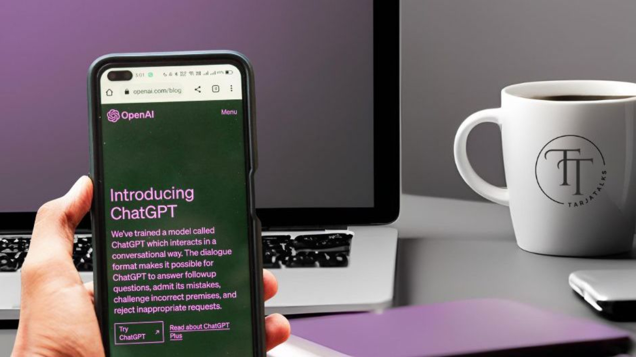 Käsi pitelee älypuhelinta, jossa ChatGPT on avoinna. Taustalla tietokone, arjen työtilanne tekoälyn käytössä.