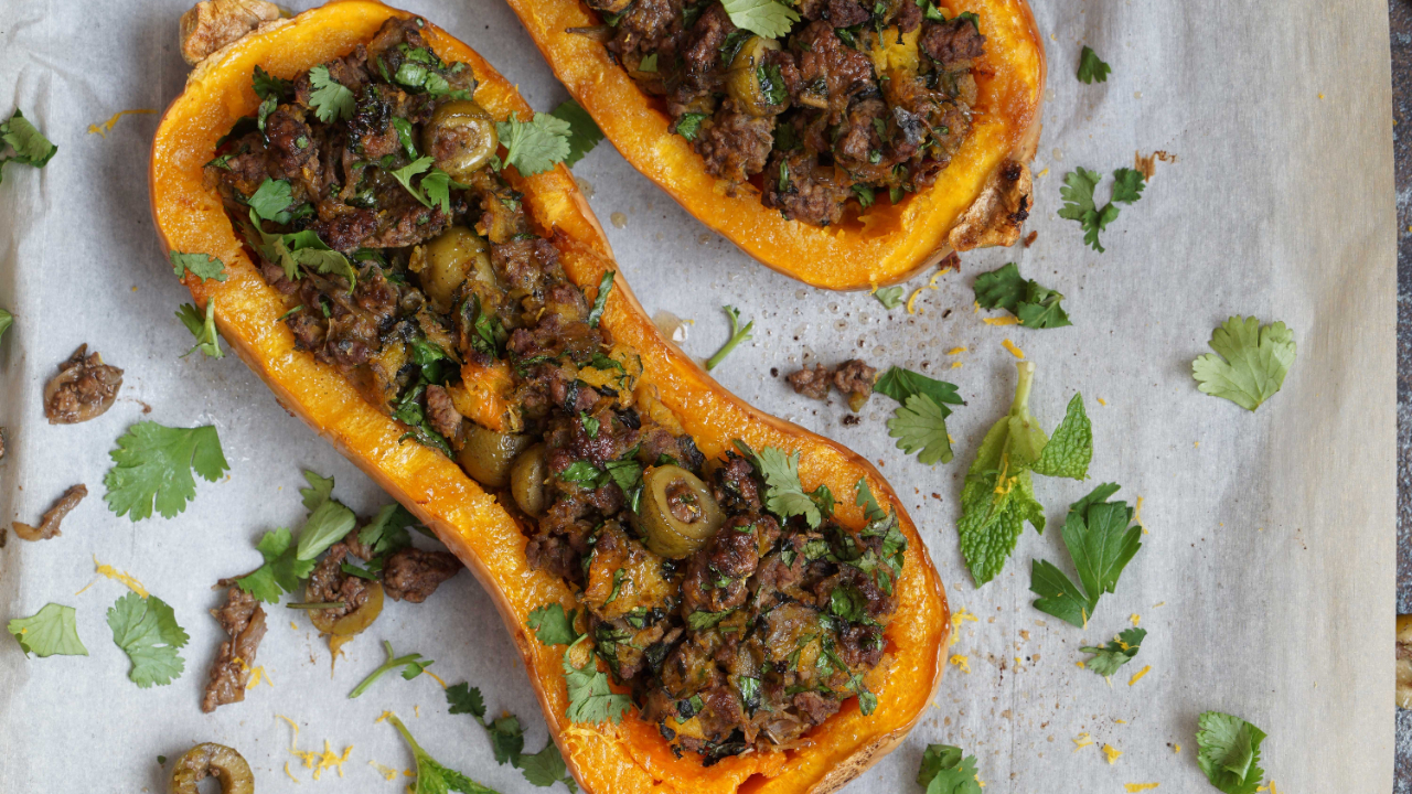 Lamb stuffed butternut boats with green olives and scattered fresh herbs like mint and parsley, on a sheet of parchment