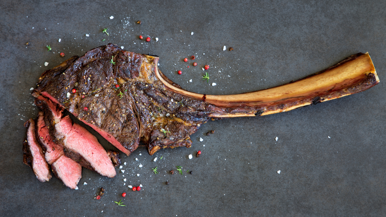 A bone-in ribeye, grilled and sprinkled with salt and pepper, sliced to reveal a juicy pink center, on a slate cutting board.