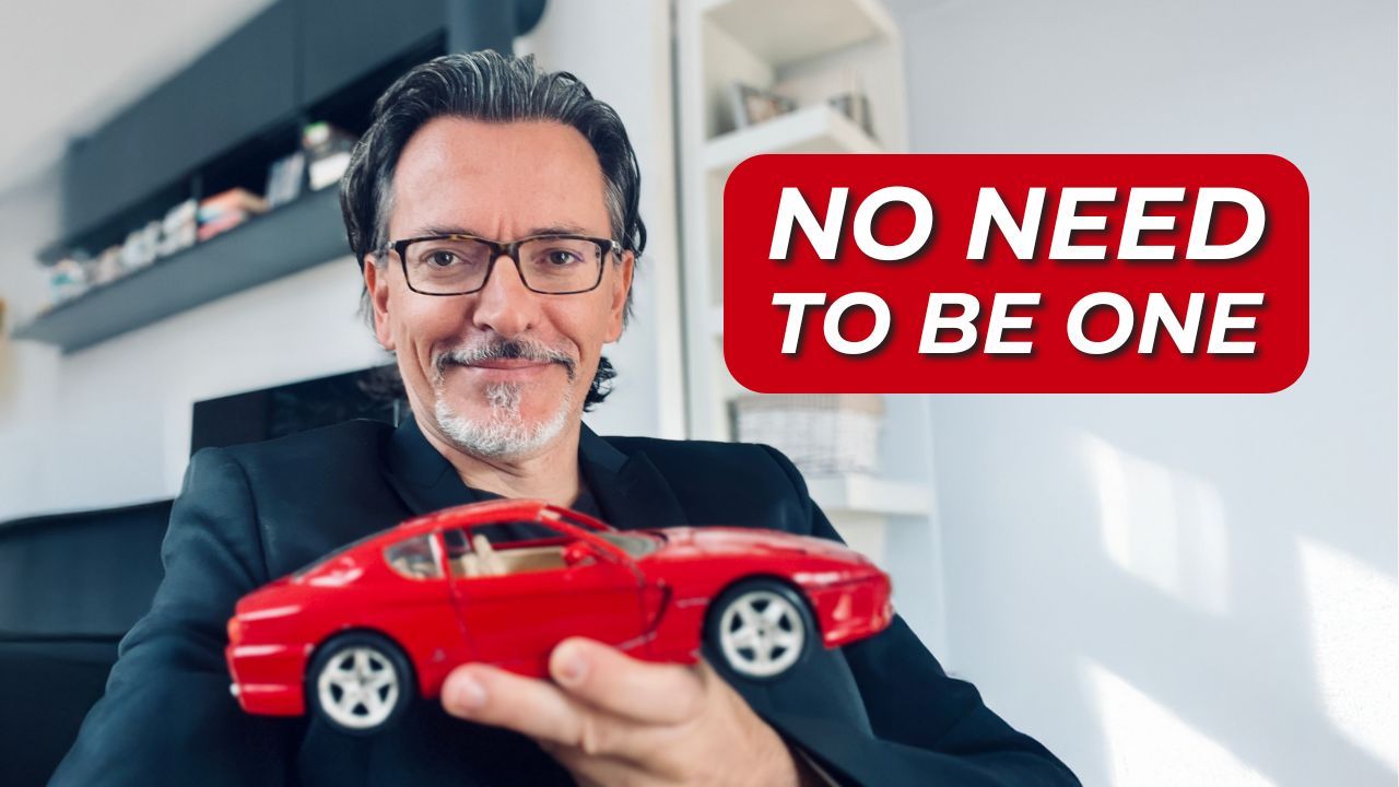 Leadership coach Gregor Prah smiling and holding a red Ferrari model car, wearing glasses and a black jacket. He is seated in a modern home office with shelves in the background. Text on the image reads: “NO NEED TO BE A FERRARI,” with “FERRARI” emphasized in red — symbolizing Gregor’s message about teamwork and leadership performance.