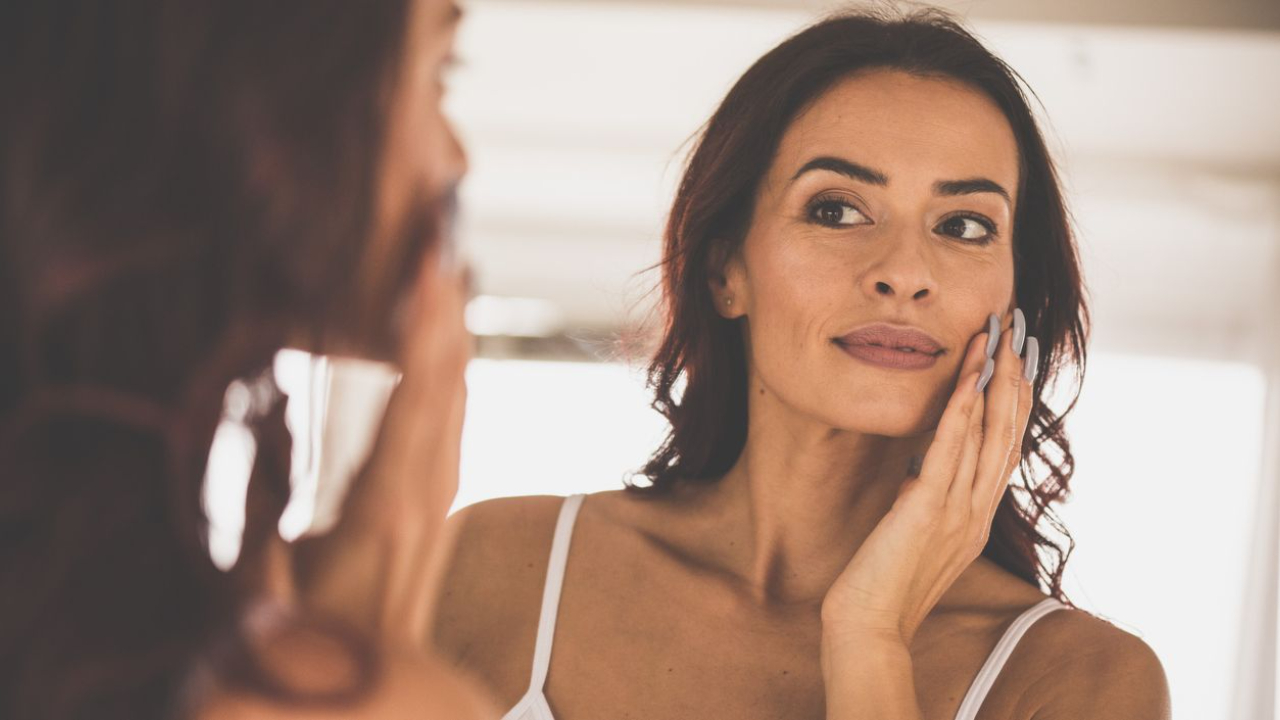 Midlife woman touching her face in the mirror, representing naturally firmer, tighter skin supported by skin tightening foods.