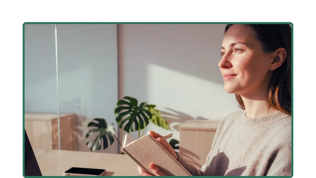 Mujer sentada junto a su escritorio con luz natural, sosteniendo una libreta antes de comenzar su día.