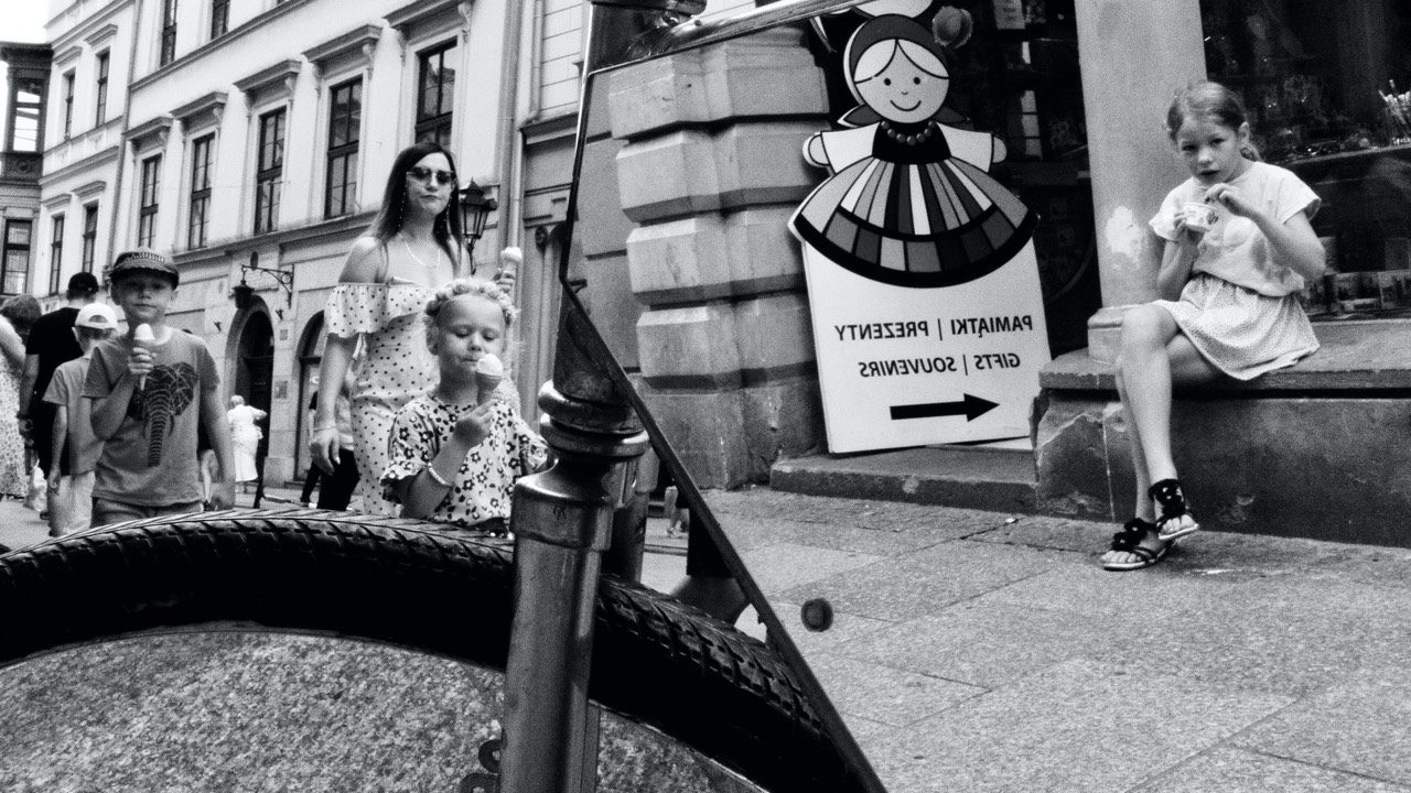 black and white street photograph of girl in mirror reflection and other girl walking behind mirror