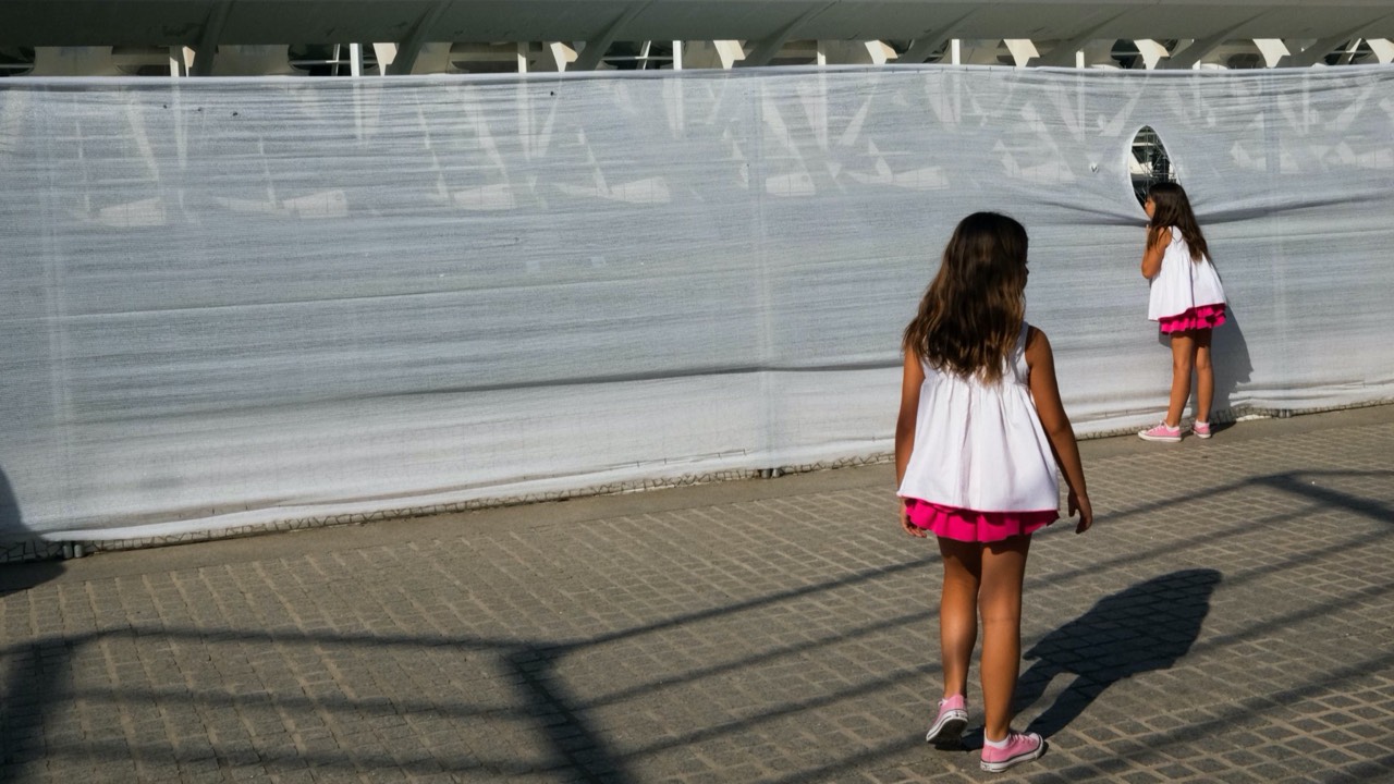 colour street photography of two girls dressed the same at the city of arts and sciences with one peeking through a hole and the other walking towards her