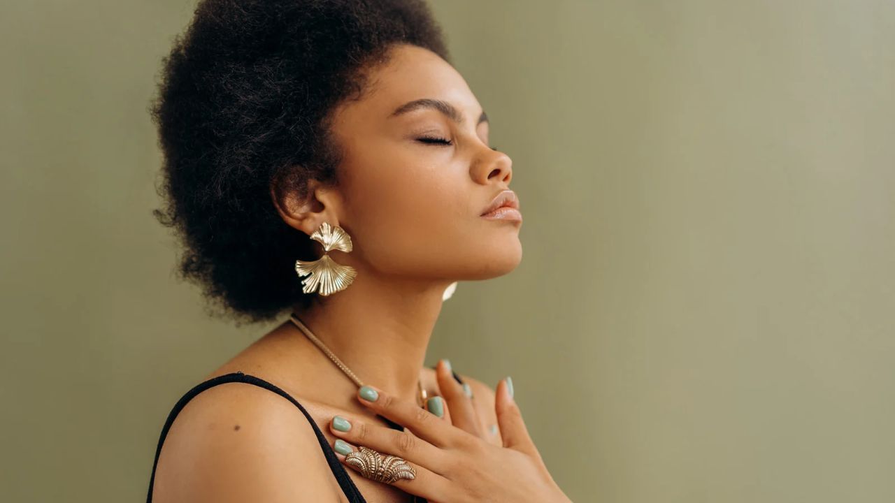 Black woman with natural hair, eyes closed, hands folded over her chest in meditation. She has short polished nails with a cuff ring on her third finger and large earrings.