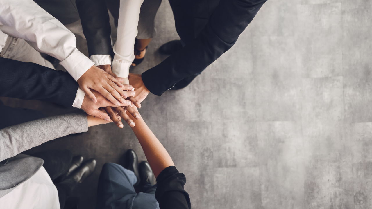 The image shows an overhead view of a group of people standing in a circle, placing their hands one on top of the other in a gesture of teamwork and unity. They are dressed in professional attire, suggesting a formal or business setting. The focus is on their hands, symbolizing collaboration and support among the team members. The floor appears to be a polished grey concrete, adding to the professional atmosphere of the setting.