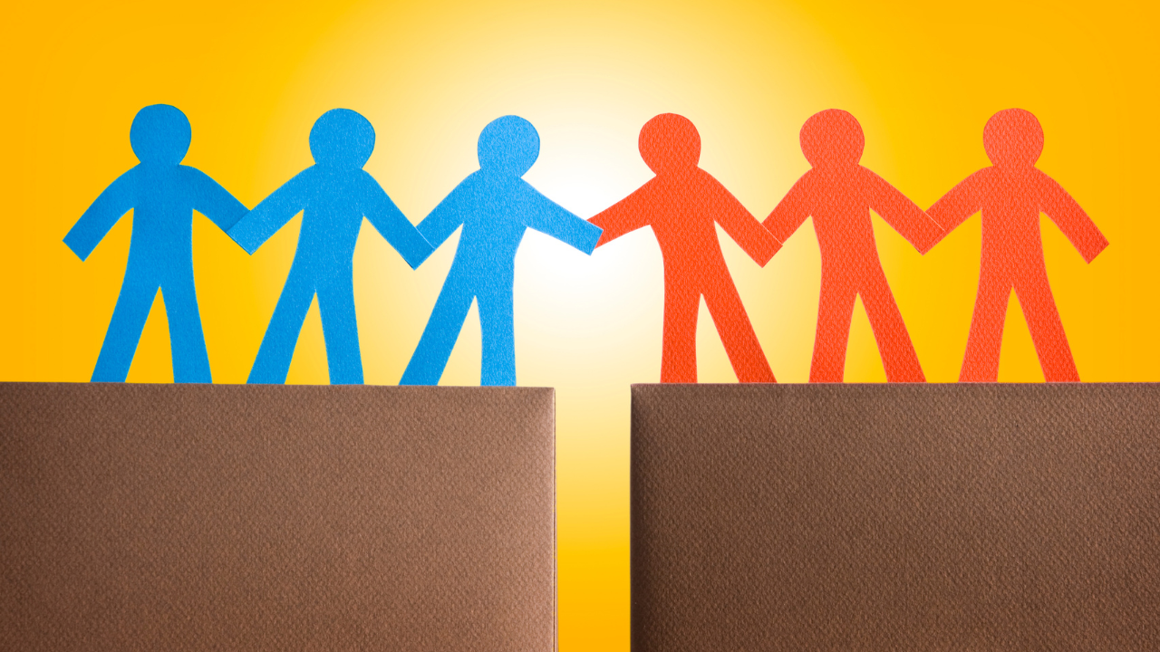 The image features a stylized representation of cooperation or partnership. It depicts paper cut-out figures in blue and red, holding hands across a gap between two brown cliffs. The background is a gradient of yellow and orange, suggesting a sunrise or sunset. This image can symbolize unity, teamwork, and the bridging of divides. The simplistic design and bright colors convey a positive and motivational message.