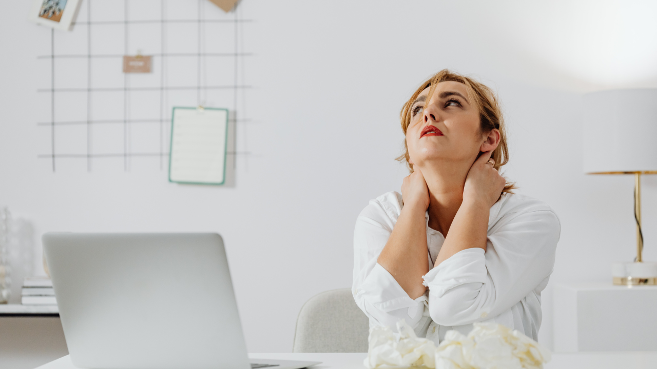 woman sitting at a desk with a laptop, holding her neck and looking upward, showing the physical strain and mental overload of overscheduling and constant work demands