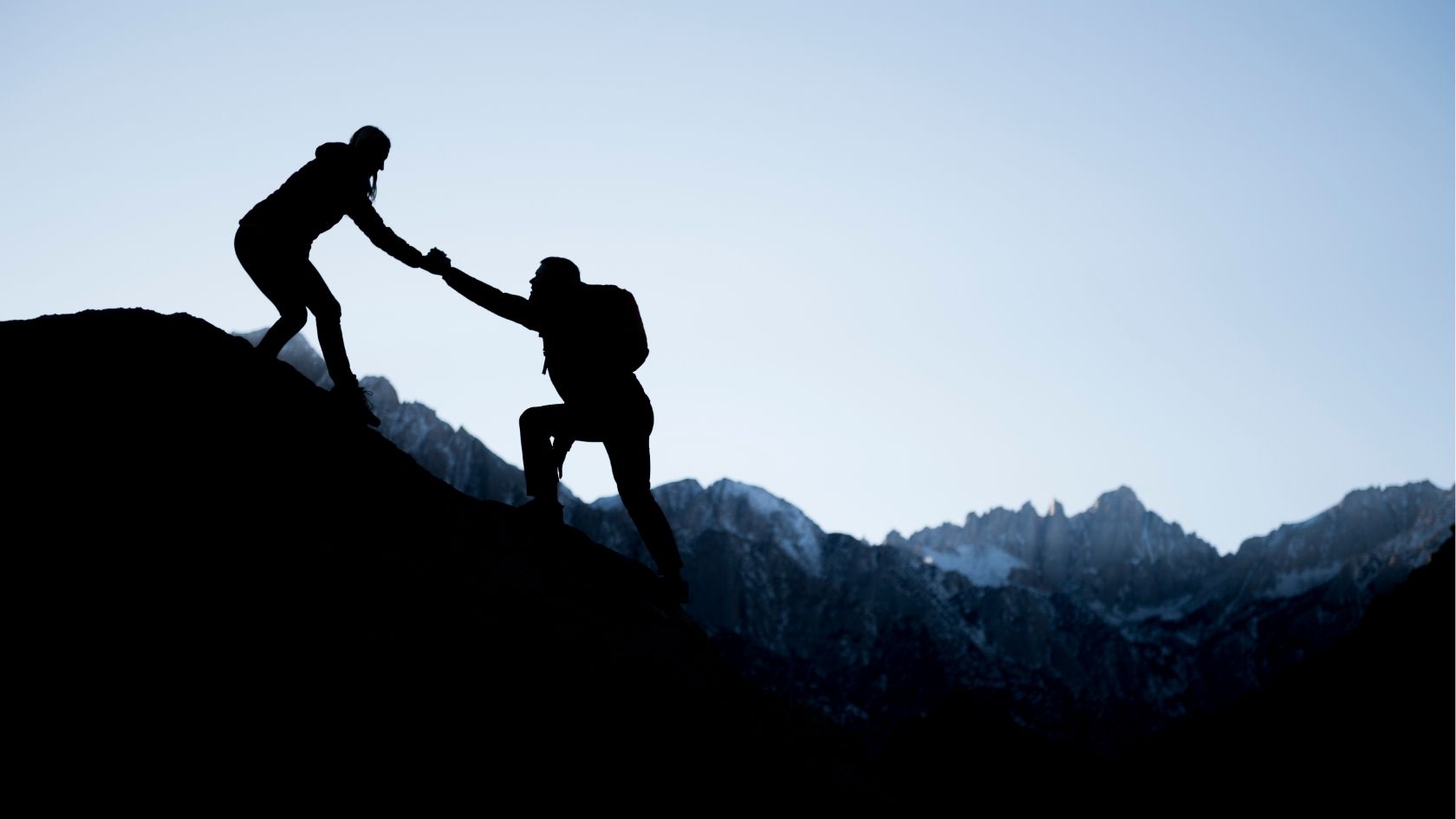 A silhouetted photo of someone helping another person up a mountain slope