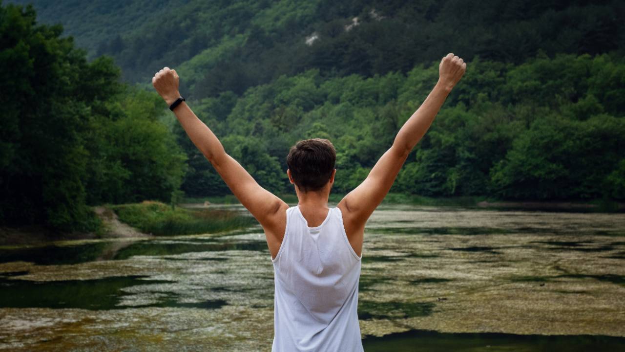 Person standing at the edge of a lake with arms raised in victory, surrounded by nature, symbolizing strength, freedom, and living with purpose after overcoming hardship.