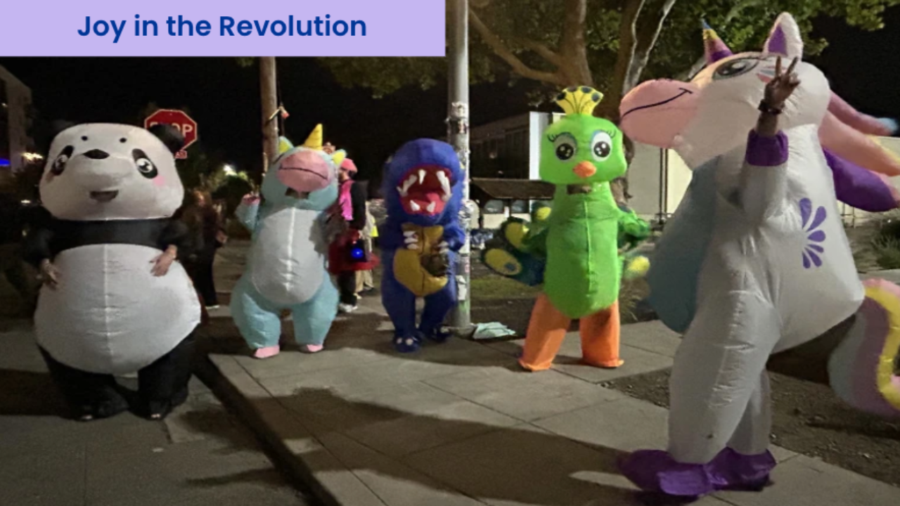 People in inflatable animal costumes, including a panda and unicorn, dancing on a city sidewalk at night beneath the words “Joy in the Revolution.”