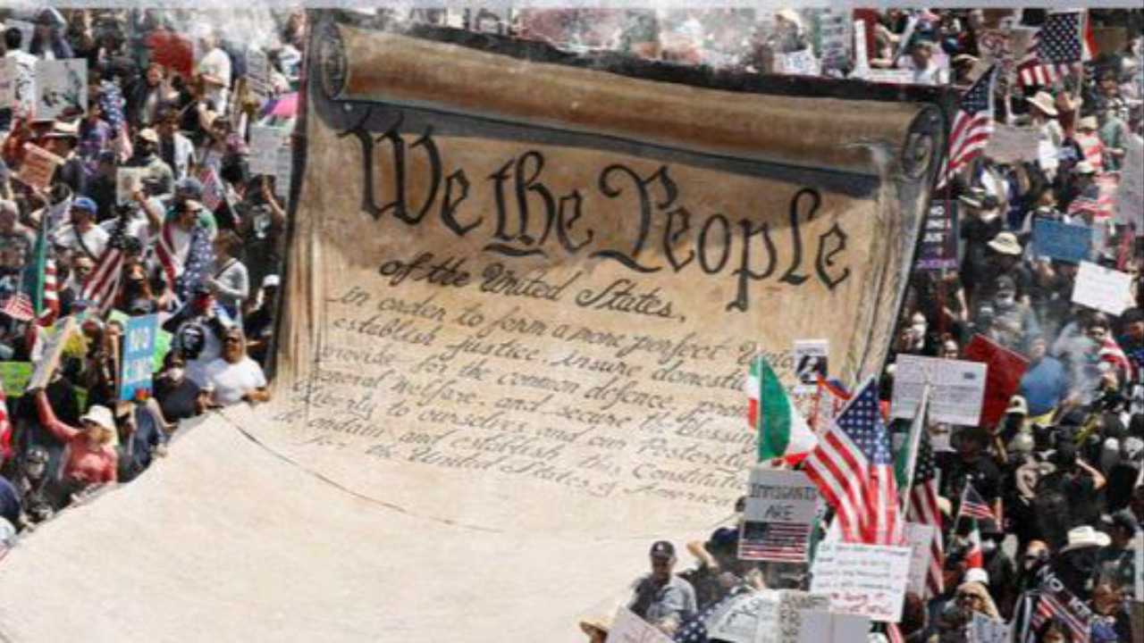A large crowd of protesters marches around a giant unfurled scroll of the U.S. Constitution reading “We the People,” holding signs and flags during a demonstration calling for protection of democratic rights.