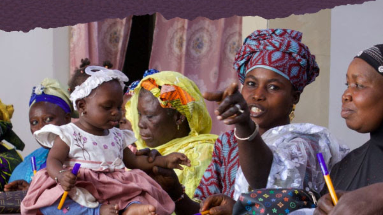 Women gathered at a table in a brightly patterned room, some holding notebooks and pens, while a baby sits on the table playing with a marker