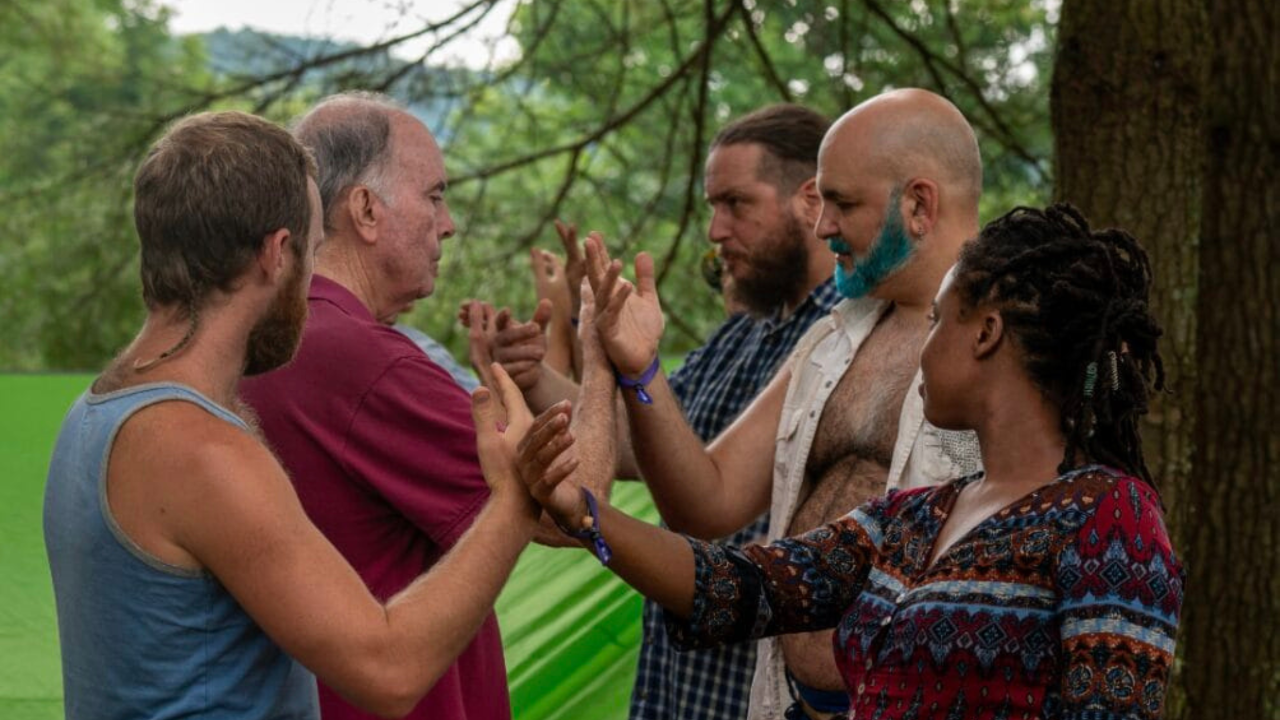 Alt text: Five adults of different ages and backgrounds stand outdoors in pairs, facing each other and gently pressing their palms together in a group trust or connection exercise among trees.