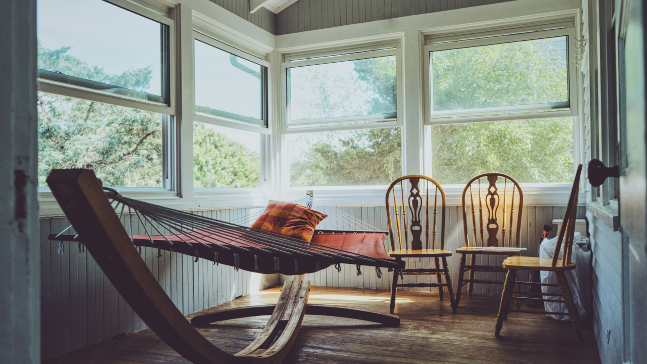 Sunlit enclosed porch with a hammock and wooden chairs, large windows overlooking green trees, creating a calm, reflective space for rest and thinking.