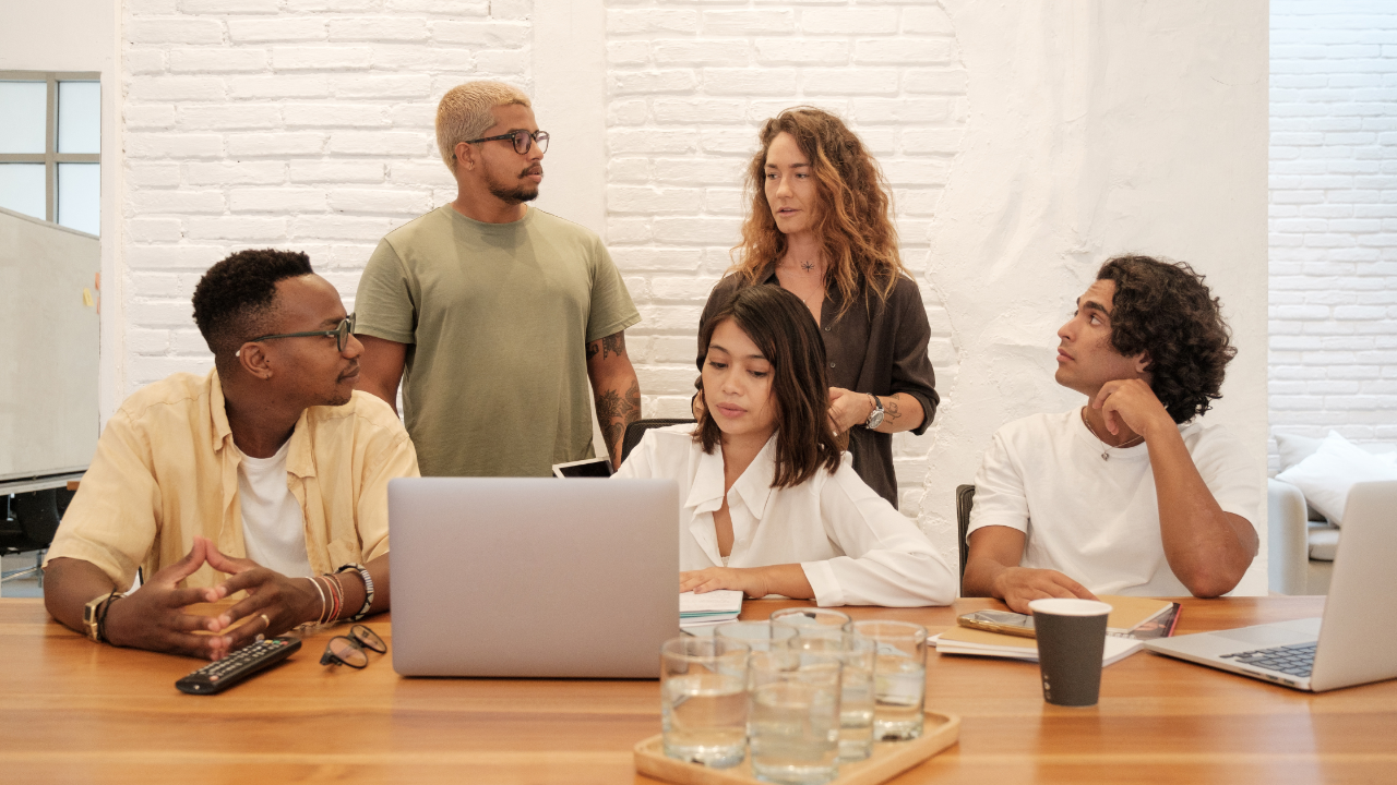 A diverse group of young adults sits around a table with laptops and notebooks, engaged in a collaborative discussion while two colleagues stand and join the conversation in a bright, modern workspace.
