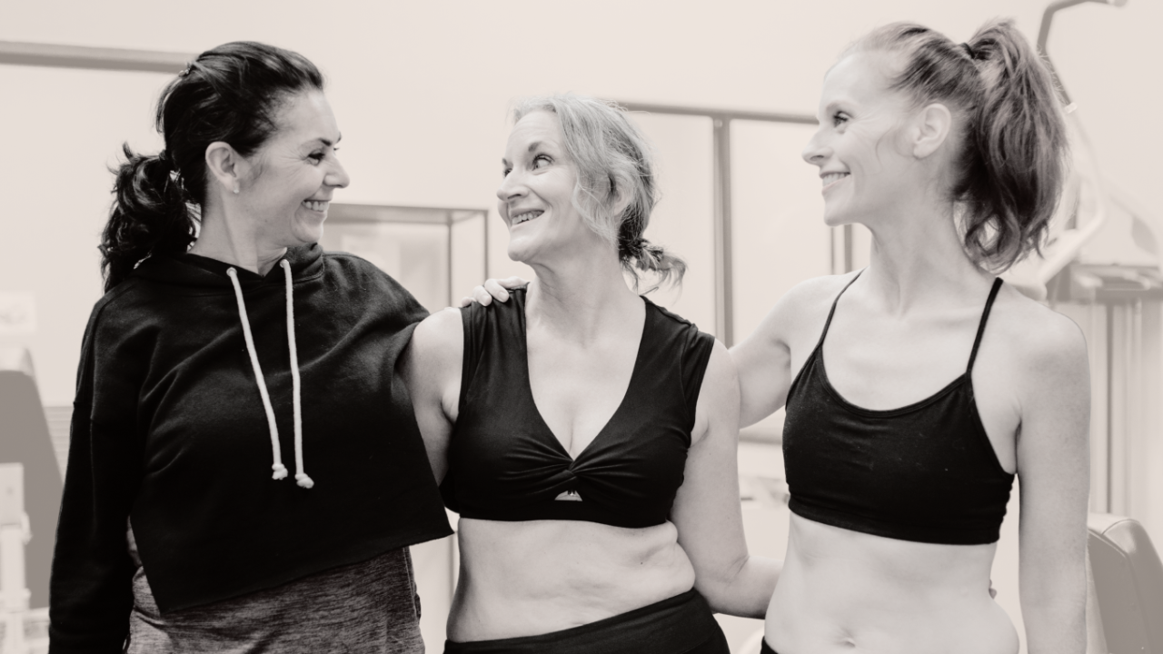 Three women stand together in a bright fitness studio, smiling and sharing a moment of laughter. They have their arms around each other’s shoulders, creating a sense of connection and support. The woman on the left wears a cropped black hoodie and ponytail, the woman in the middle wears a black sports bra with a small knot detail, and the woman on the right wears a black spaghetti-strap workout top with her hair tied back. The photo is in black and white, emphasizing warmth, authenticity, and friendship among women in midlife.