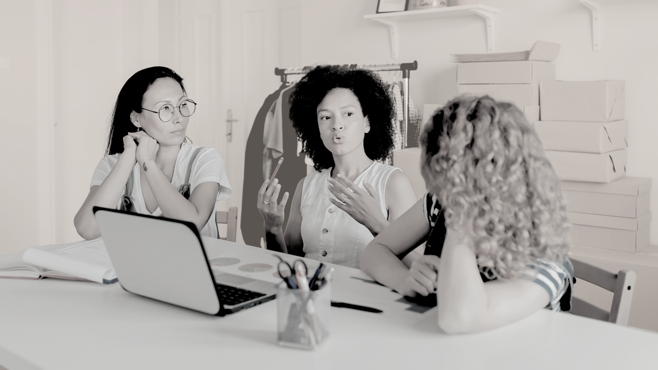 Three women sit around a white table in a small creative studio space. One woman with curly hair is speaking with her hands, mid-explanation, while the other two listen closely. A laptop, notebooks, and pens are spread out on the table. Clothing racks and shipping boxes line the background, giving the room a working, entrepreneurial feel. The color tone is muted and soft, creating a grounded, collaborative atmosphere.