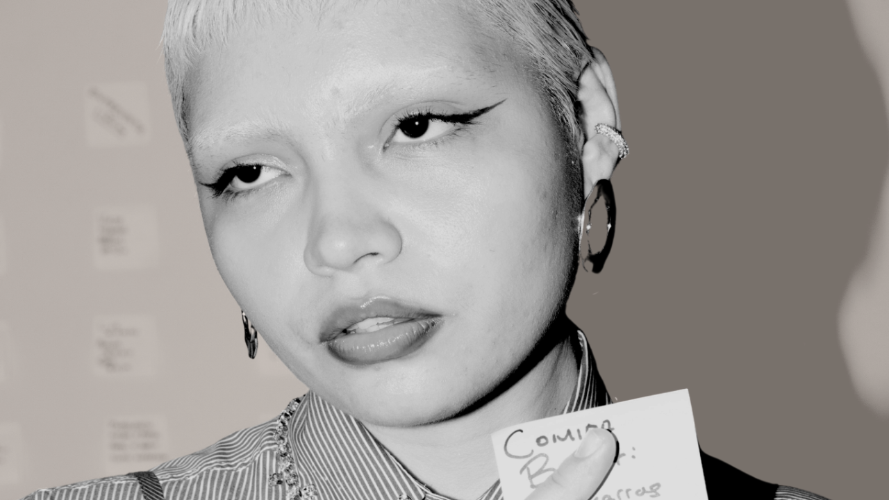 A close-up black and white portrait of a person with bleached short hair, minimal eyebrows, winged eyeliner, and glossy lips. They wear hoop earrings and a striped shirt with layered necklaces. Their expression is calm and introspective as they hold a small notecard in one hand. The background is softly blurred with indistinct shapes and notes visible.