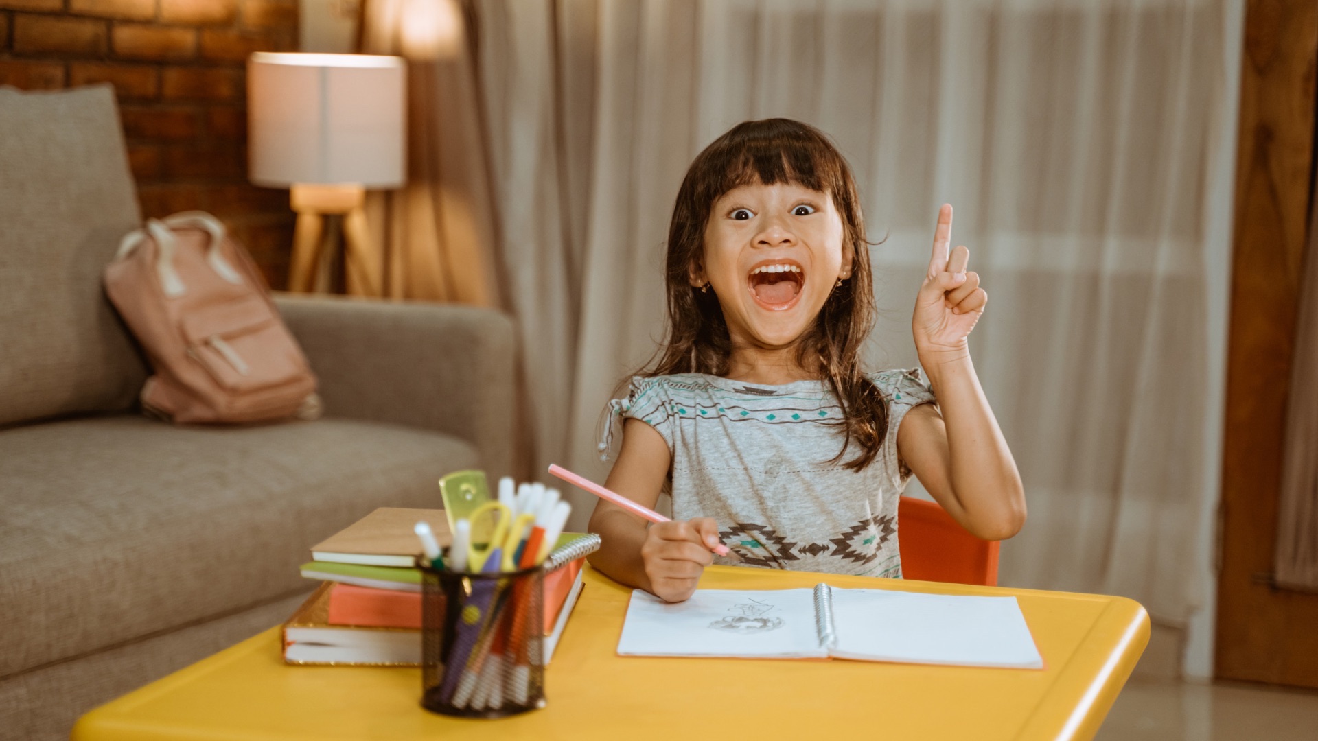 A smiling young girl excitedly learning at home, holding up her finger with an idea during a homeschool lesson.