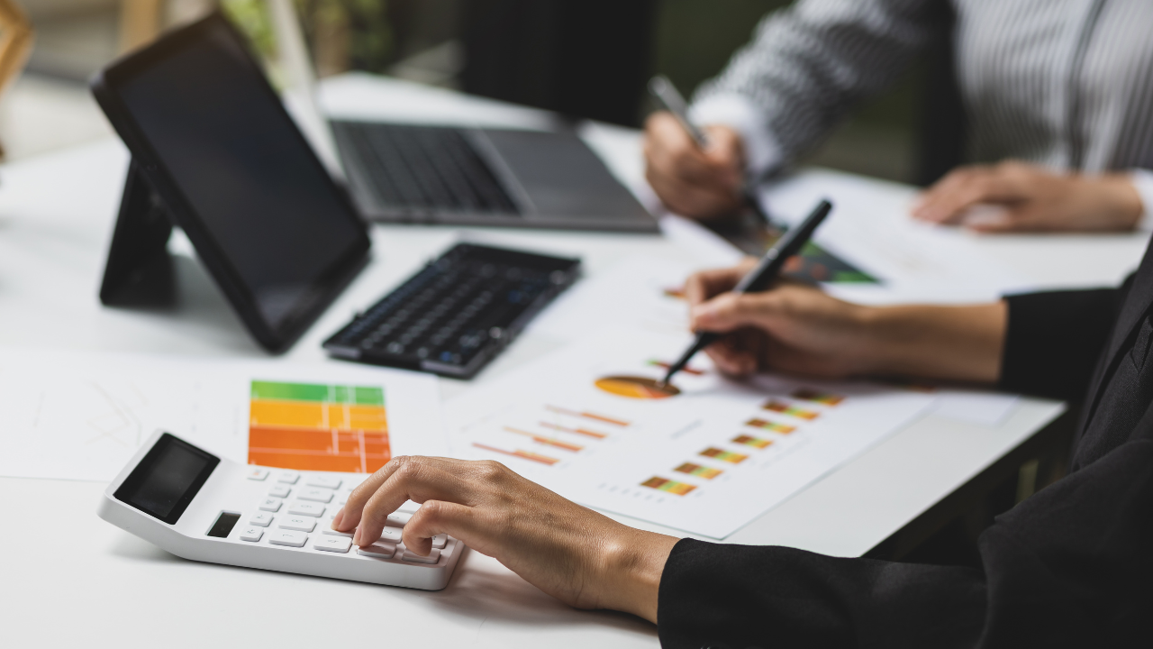 Image of a desk with a calculator, electronic device and someone using the calculator and writing on a graph.  The image is of the hands only.