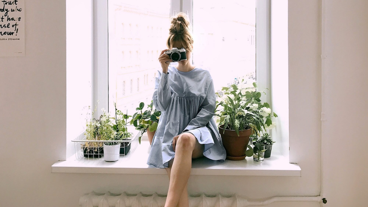 a female sitting on a windowsill holding a camera up to her eye - surrounded by plants.