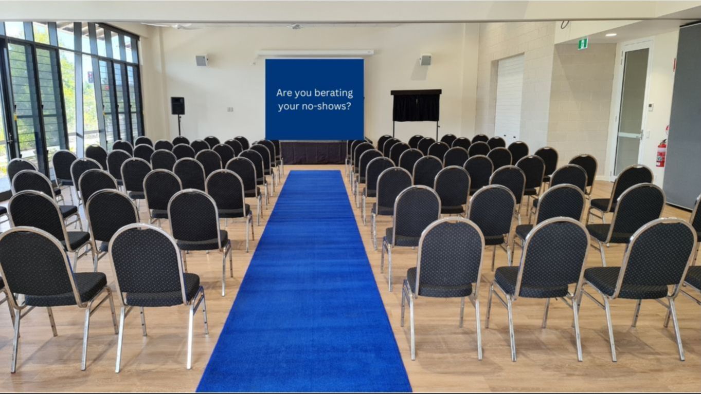 Photo of a room with an event set theatre style with rows of black chairs and a blue carpet down the middle. The perspective is from the back of the room, looking down the blue carpet to the stage.
