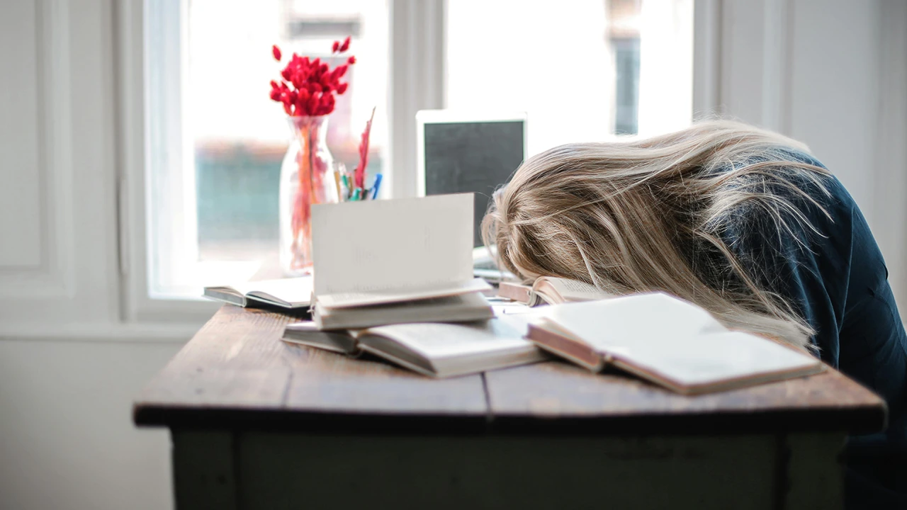Exhausted woman with head on desk surrounded by books and laptop, showing signs of fatigue from menopause and thyroid imbalance
