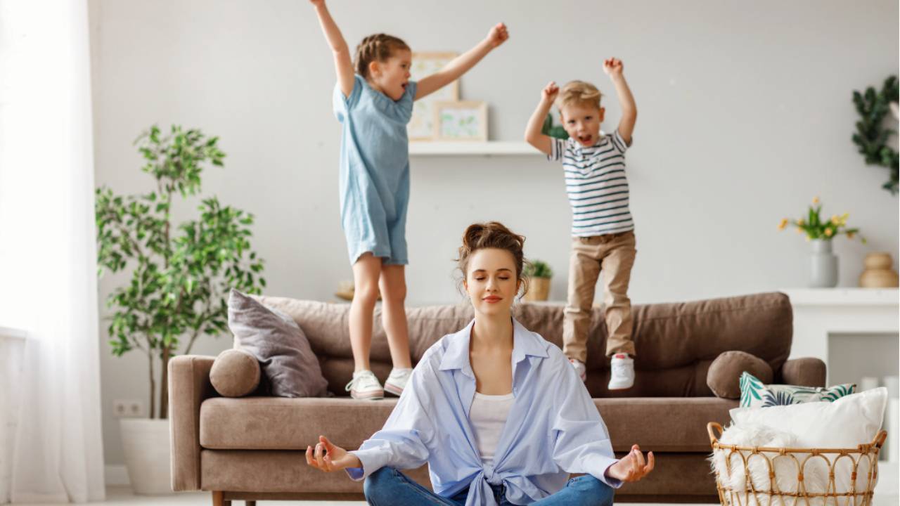 Woman meditating on the floor while children jump on the sofa behind her