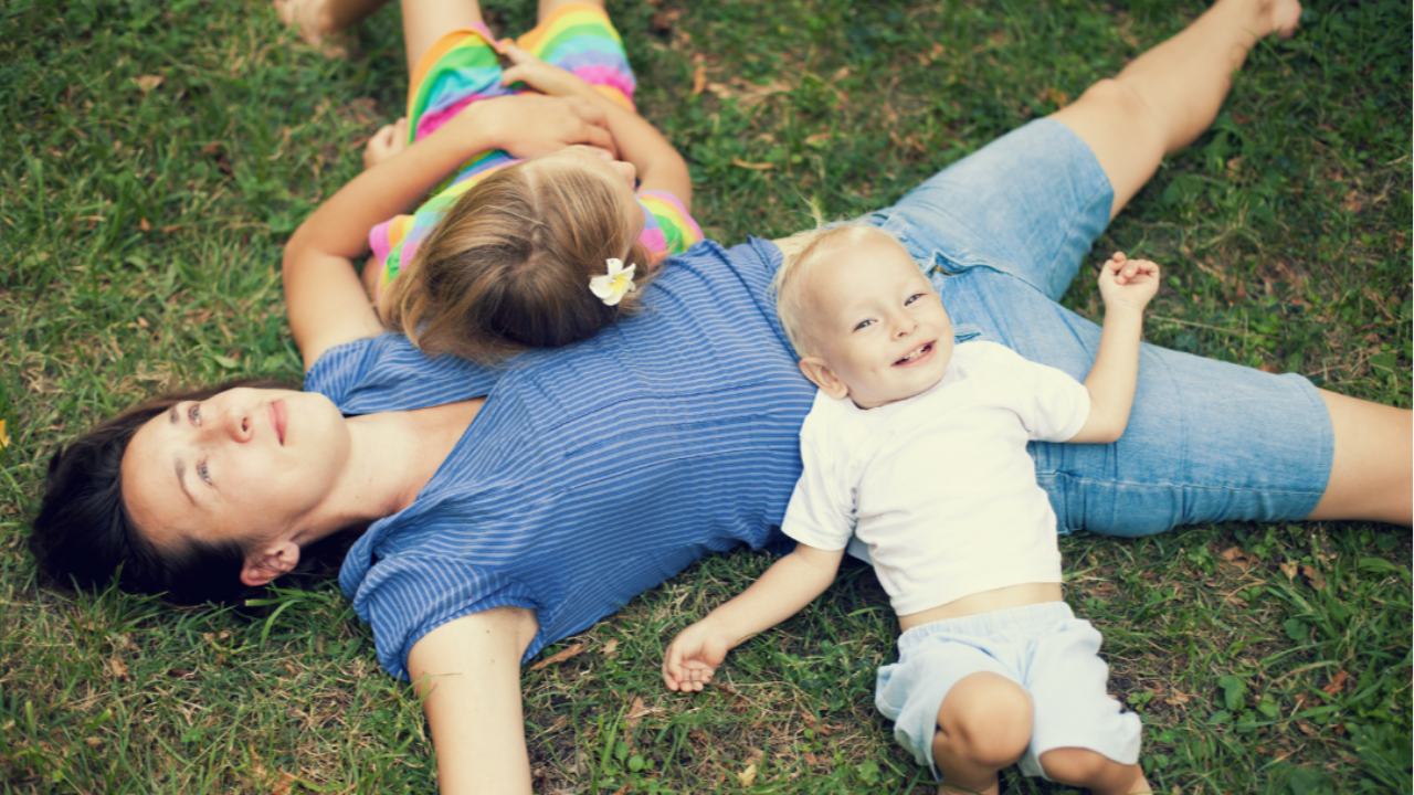 Mother lying in grass enjoying time with her two young children, smiling and relaxed outdoors