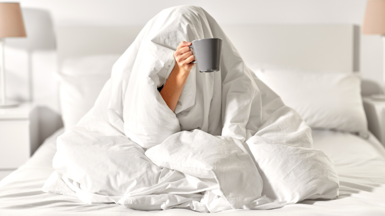 Woman hiding under a duvet, hand poking out clutching a mug