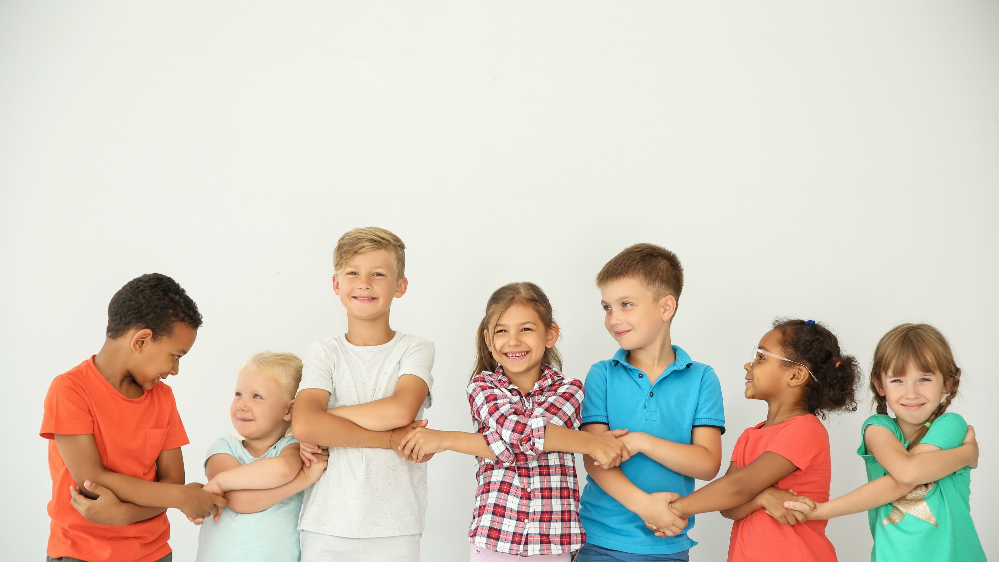 A smiling boy with curly hair stands in a classroom with his finger pointed up, suggesting he has a great idea. Three other children sit in the background, near a bookcase. One boy is standing up, and two girls are seated. The classroom has light walls, and natural light fills the room. This photo could be used in projects about education, schools, children, or learning.
