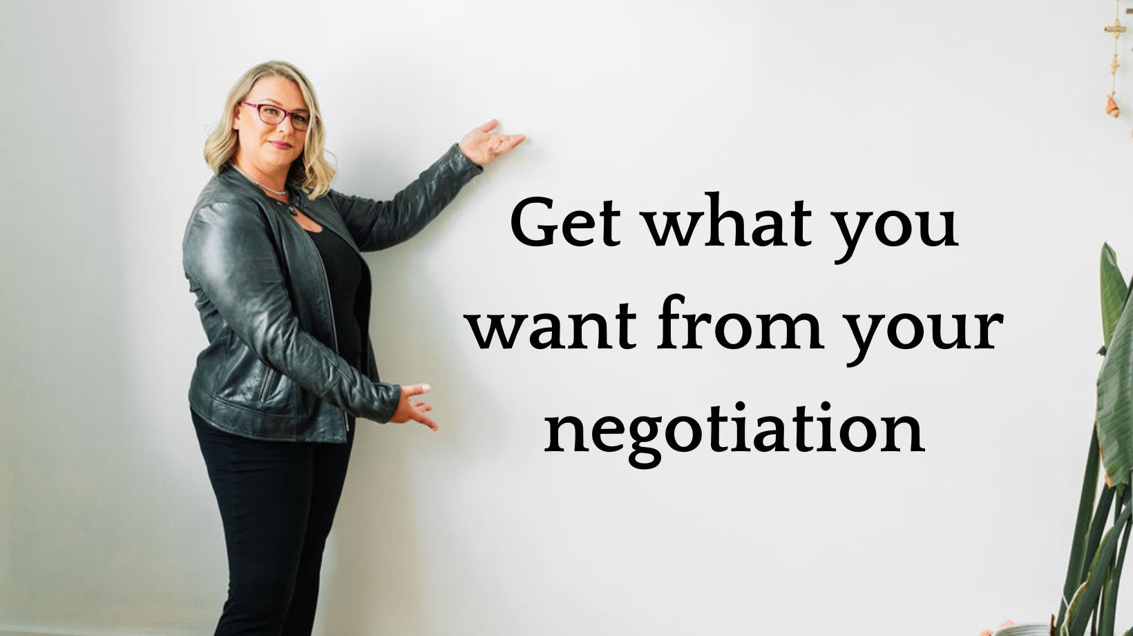 Joanne is standing in front of a white wall and gesturing to text which reads 'Get what you want from your negotiation'. She appears warm and confident, and ready to help you learn what you want from a negotiation.