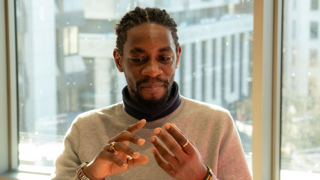 black man with hair in braids, looking at his fingers whilst thinking