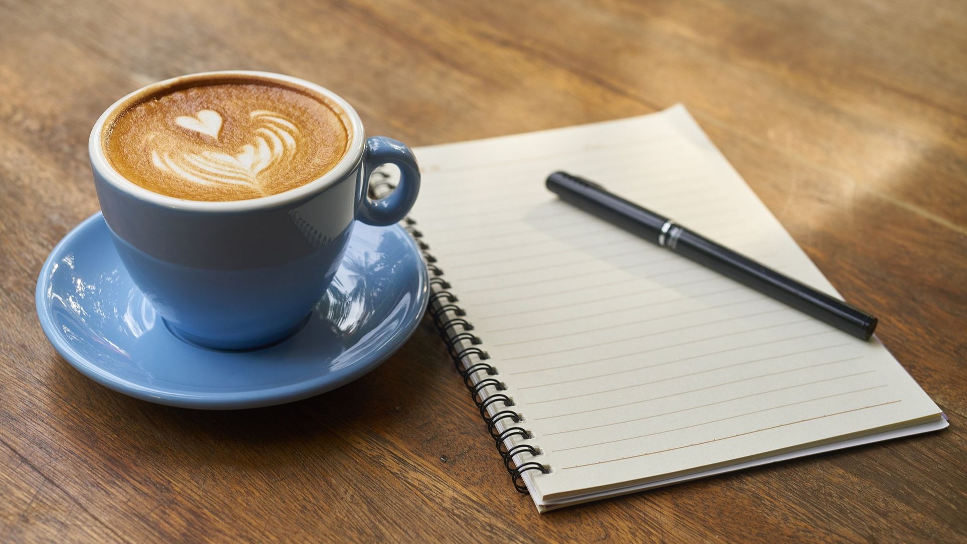 A latte in a blue cup and saucer sits on a table next to a journal by Engin Akyurt of Pixabay