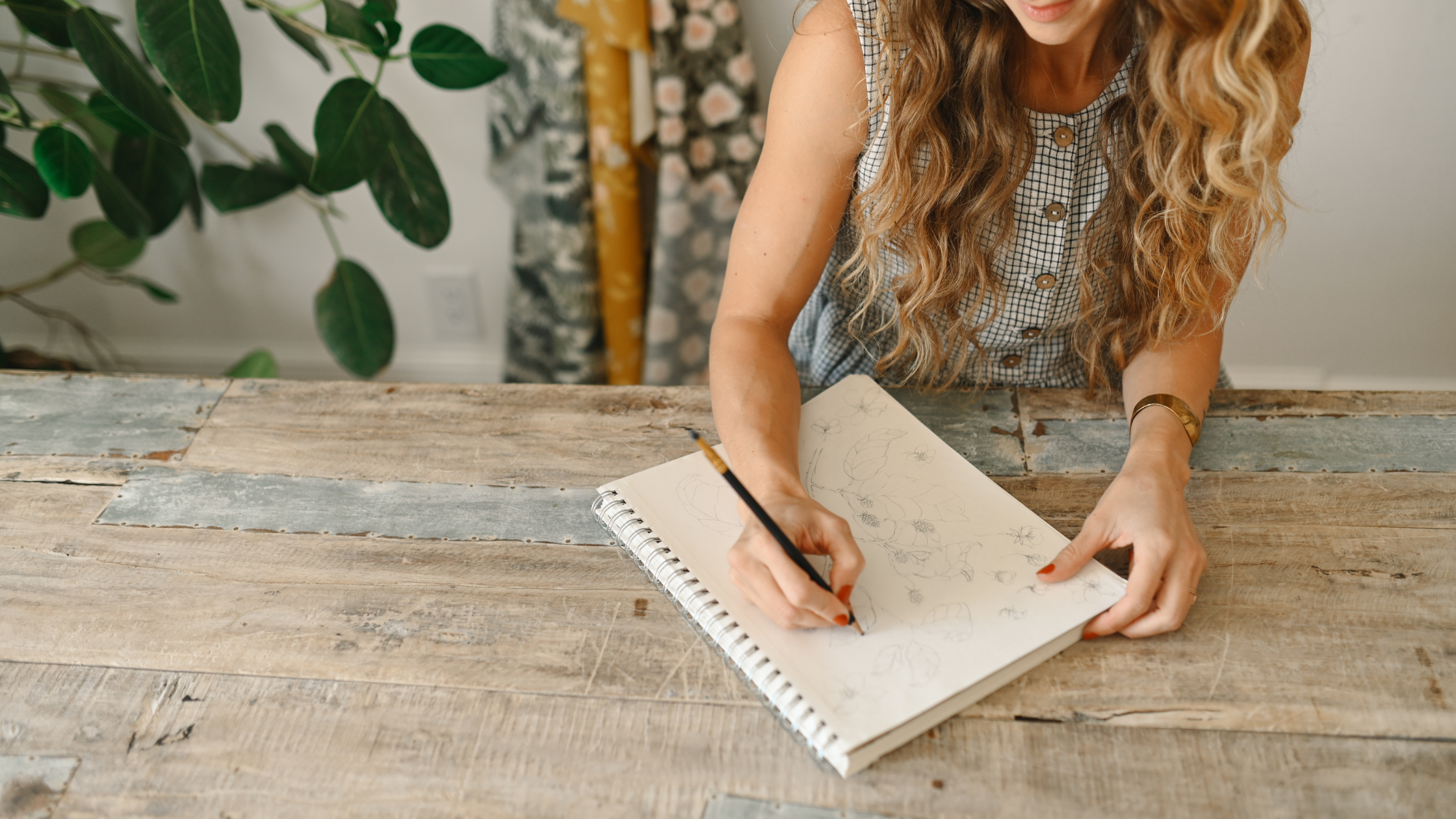 Surface Pattern Designer and Creative Entrepreneur Bonnie Christine sits at a rustic wooden table, sketching delicate floral patterns in a notebook. Her thoughtful expression and creative process reflect her passion for design and artistry.