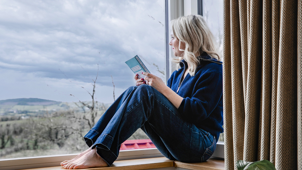 Image of Sinead Brady sitting with her barefoot with her feet on a window sill, looking out over the countryside while holding her book Total Reset in her hand.  