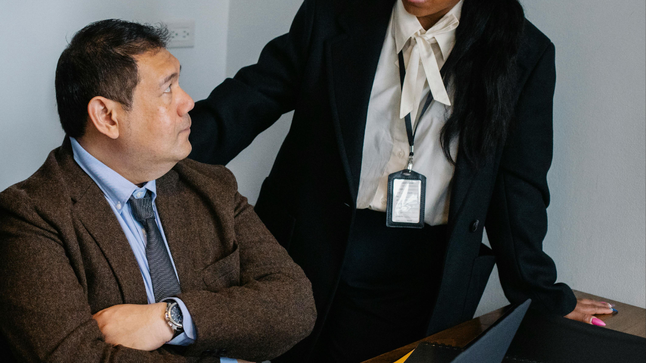 A pre-retirement employee sitting in front a laptop with his arms crossed in front of him looking up at a woman in a suit and looking disengaged.