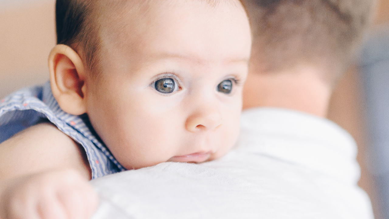 Awake baby resting on dad’s shoulder, looking alert but unsettled, representing sleep training readiness concerns