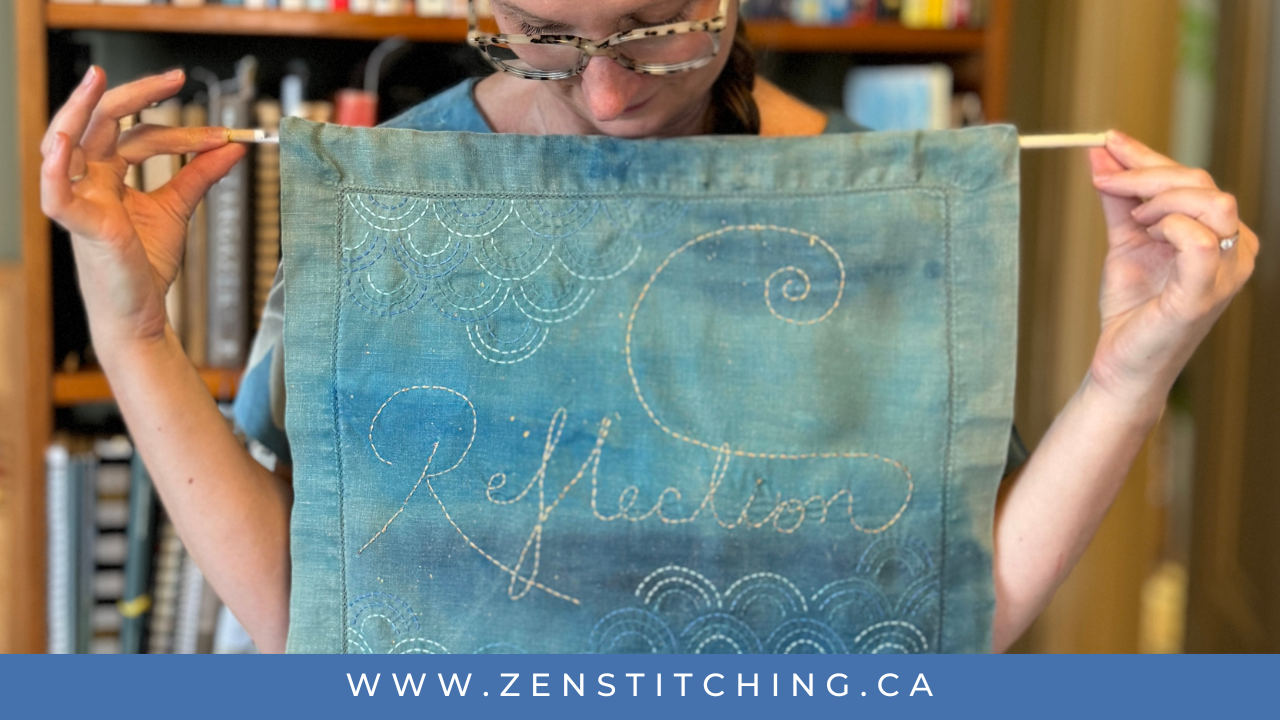 A white woman holds a piece of stitching towards the camera whilst looking down at it. The piece says 