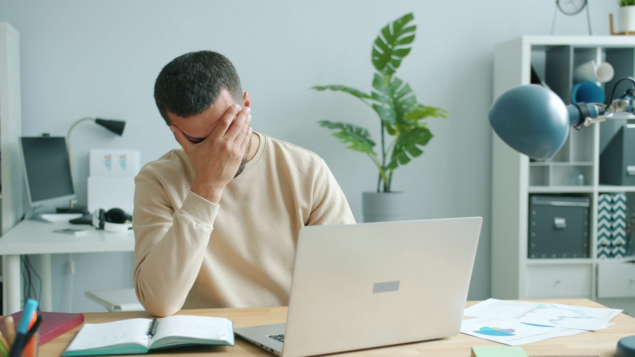 Exhausted man covering his face with his hand while sitting at a desk in front of an open laptop in a bright home office
