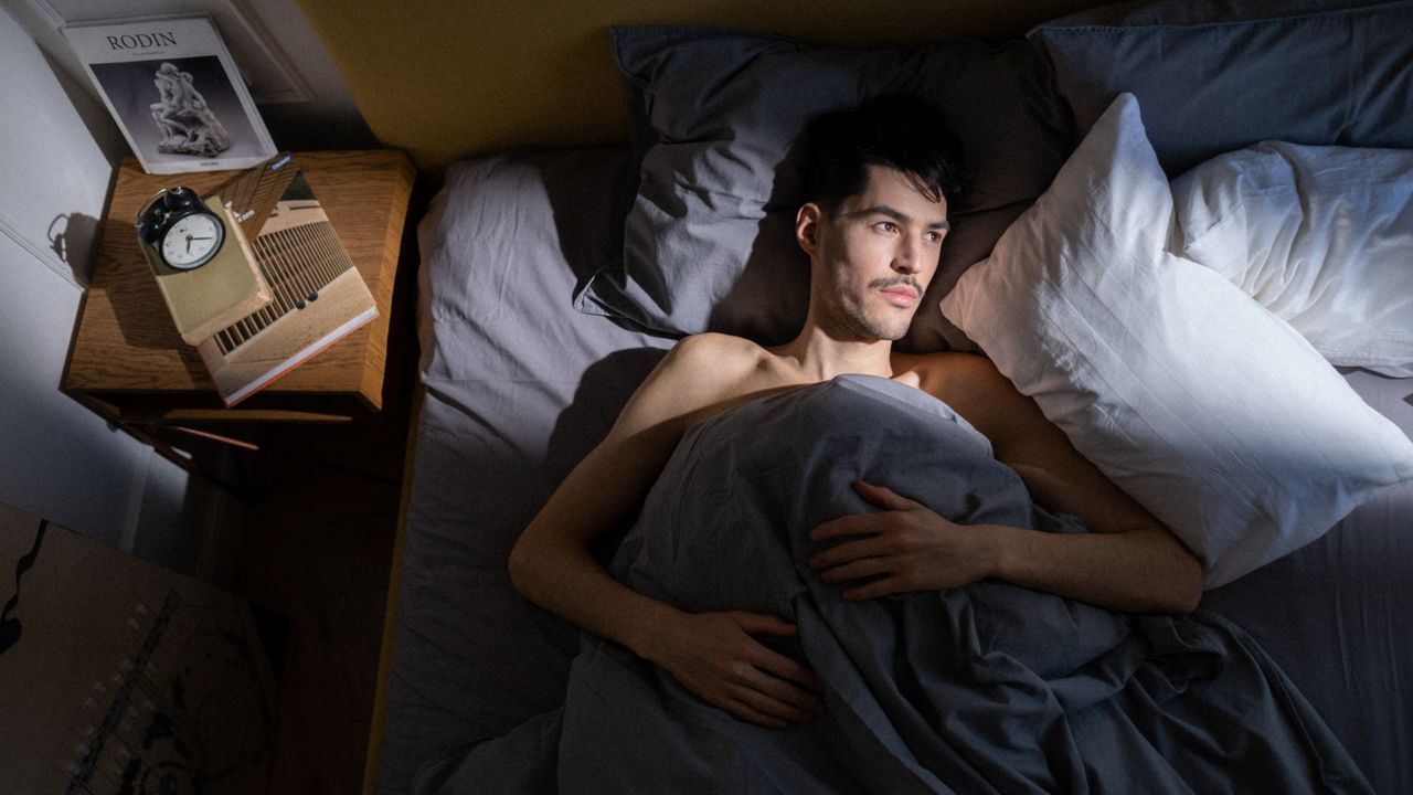 Man lying awake in bed beside a bedside table with an alarm clock and books