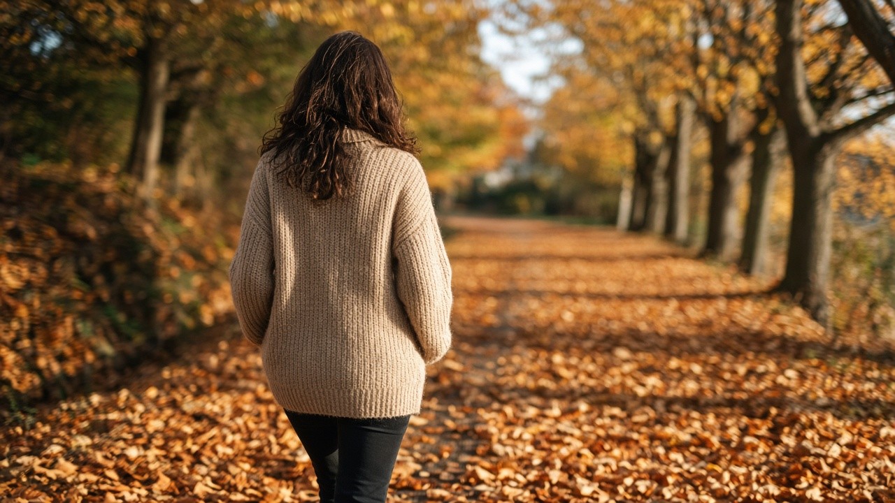 Woman in a beige knit jumper walking along a leaf covered autumn path on a sunny morning
